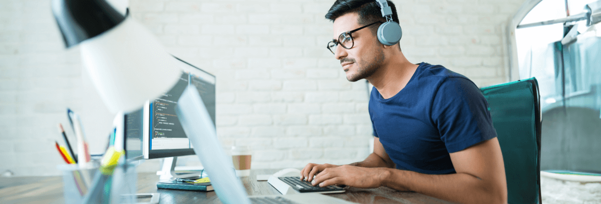 A person wearing headphones is focused on working at a laptop in a casual office setting, with a notepad, pen, and smartphone on the desk and a blurred indoor background.